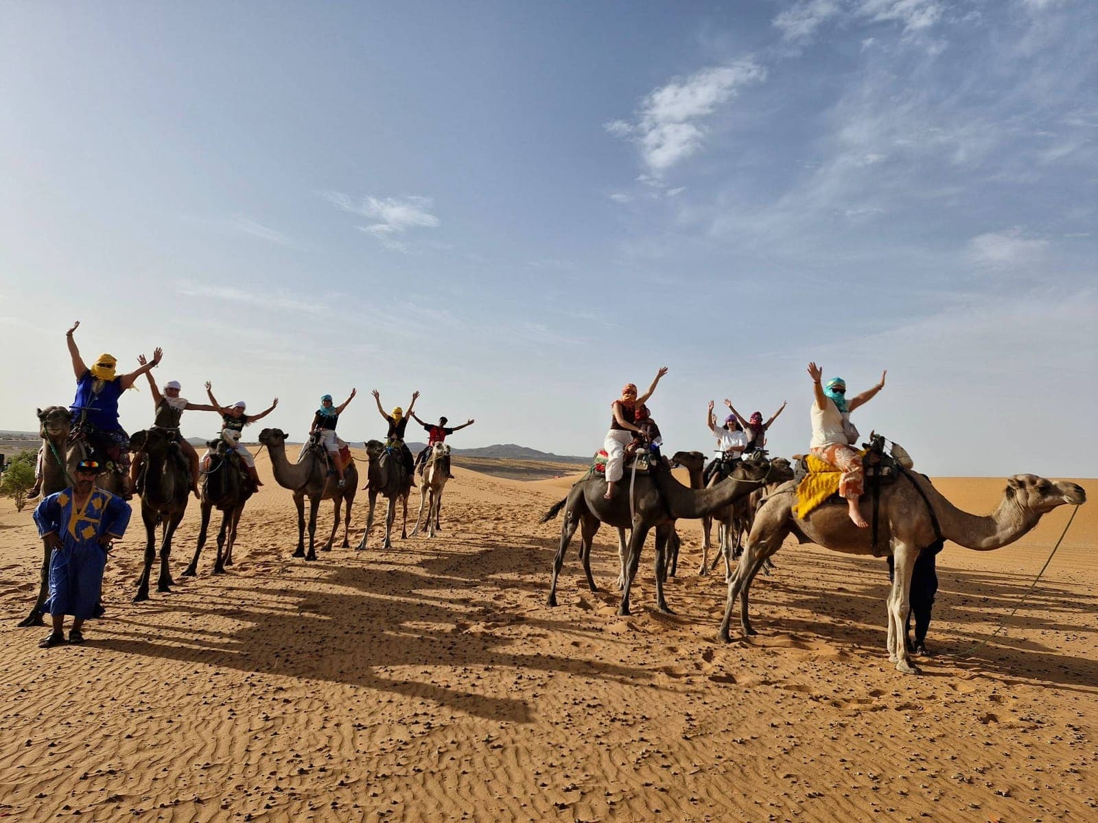 A group of travellers on dromedaries waving with raised arms in the desert dunes, beside a guide in a blue robe