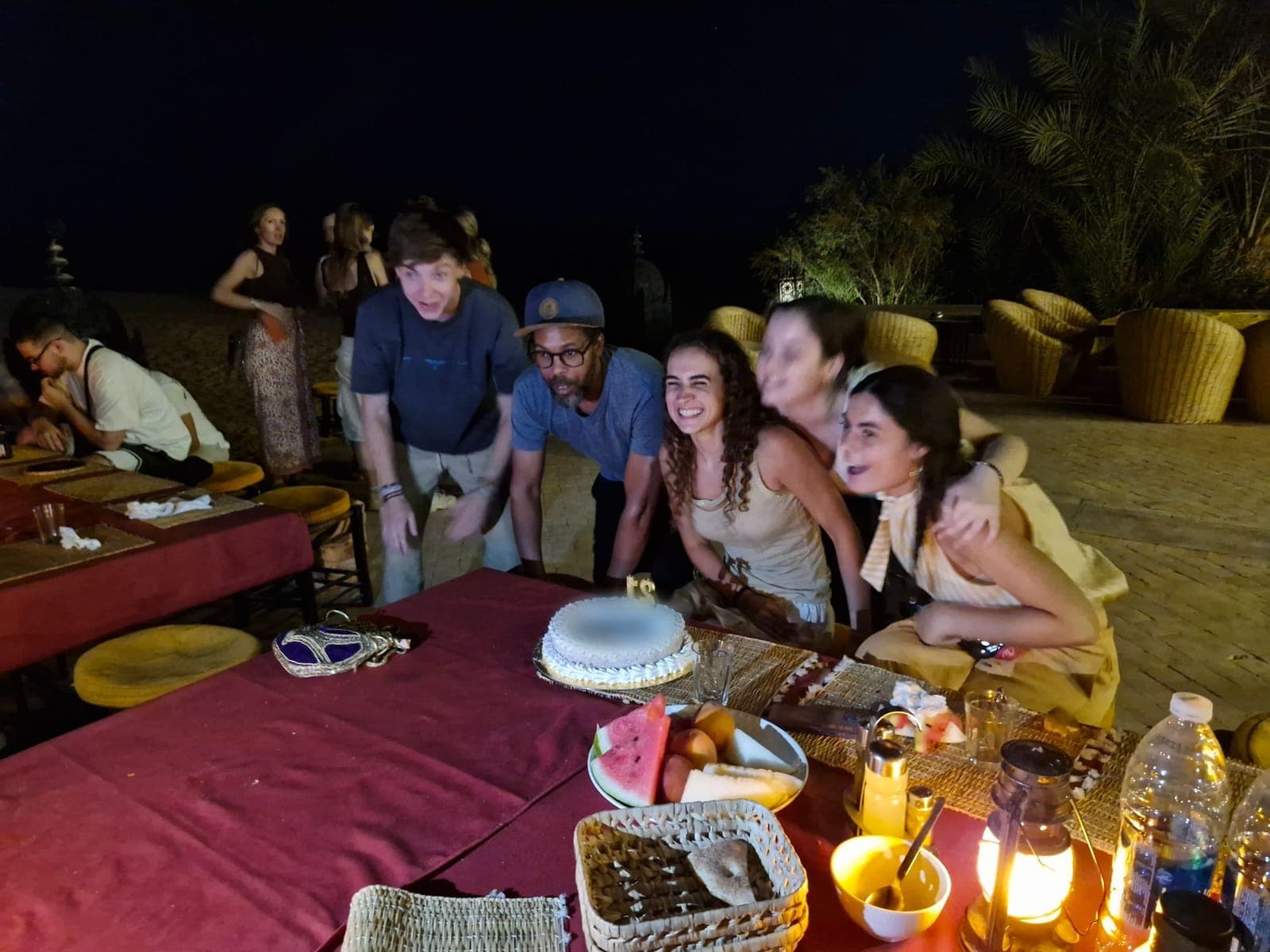 Travellers celebrating with a cake at an open-air dinner at night, with lanterns on the table